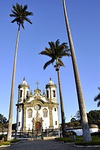 Igreja São Francisco de Assis - Foto da Igreja São Francisco de Assis. Na frente, 3 palmeira muito altas, atrás das pameiras, uma igreja com duas torres paralelas na fachada, detalhes barrocos em pedra sabão. Porta grande vermelha no centro da fachada da igreja. Há alguns arbustos dispostos ao redor e na frente da igreja. Ao fundo o céu azul claro sem nuvens.