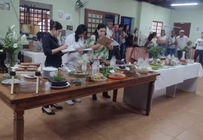 Mesa com pratos diversos sendo avaliado por três mulheres e algumas pessoas ao fundo assistindo