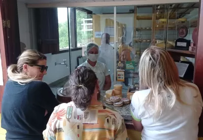 Mulher de máscara e touca apresenta queijos artesanais a três clientes