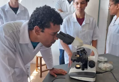 Estudante de jaleco utiliza um microscópio com outros colegas ao redor