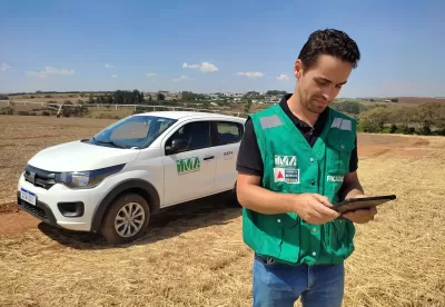 técnico do instituto mineiro de agropecuária veste um colete da instituição, de pé, segurando um tablet com um carro do IMA ao fundo em um campo aberto
