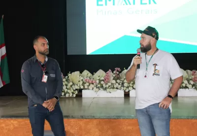 Dois homens se apresentam em um evento, o da esquerda está com um microfone nas mãos e camisa da Epamig