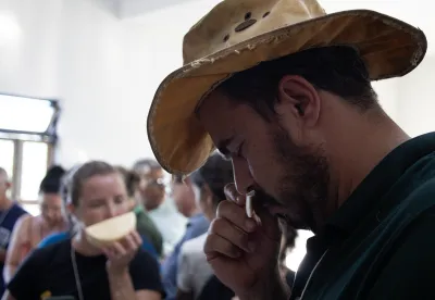 Foto de homem com um chapéu sentindo o aroma de um pedaço de queijo. Ele segura o queijo na mão e está com os olhos fechados.