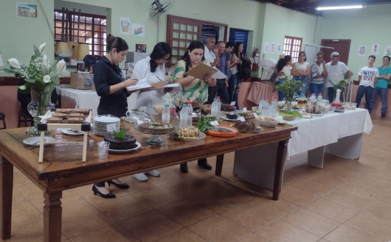 Mesa com pratos diversos sendo avaliado por três mulheres e algumas pessoas ao fundo assistindo