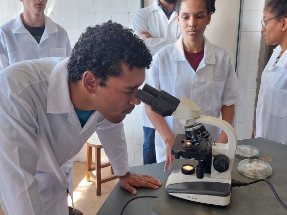 Estudante de jaleco utiliza um microscópio com outros colegas ao redor