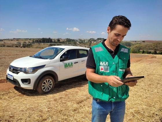 técnico do instituto mineiro de agropecuária veste um colete da instituição, de pé, segurando um tablet com um carro do IMA ao fundo em um campo aberto