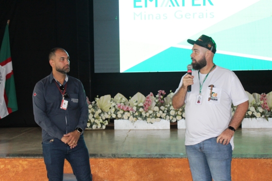 Dois homens se apresentam em um evento, o da esquerda está com um microfone nas mãos e camisa da Epamig