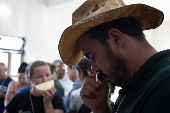 Foto de homem com um chapéu sentindo o aroma de um pedaço de queijo. Ele segura o queijo na mão e está com os olhos fechados. 