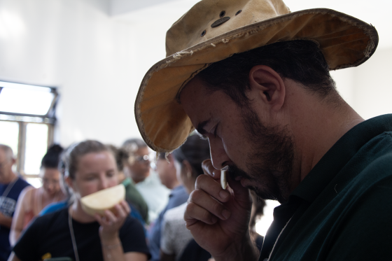 Foto de homem com um chapéu sentindo o aroma de um pedaço de queijo. Ele segura o queijo na mão e está com os olhos fechados.