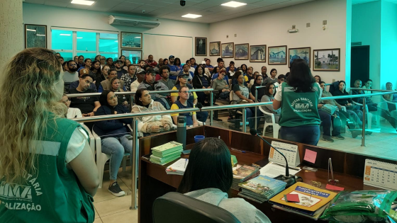 Mulheres com uniformes escrito Defesa Sanitária IMA diante de plateia. Uma deles de costas fala para a plateia.